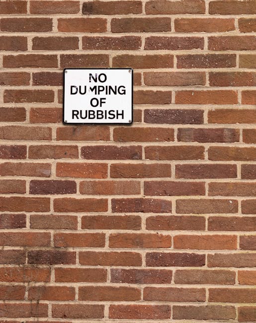 A large red metal dumpster with visible rust and wear is positioned against a dark green wall and a light gray concrete wall, situated on a paved outdoor surface. The dumpster's surface shows signs of