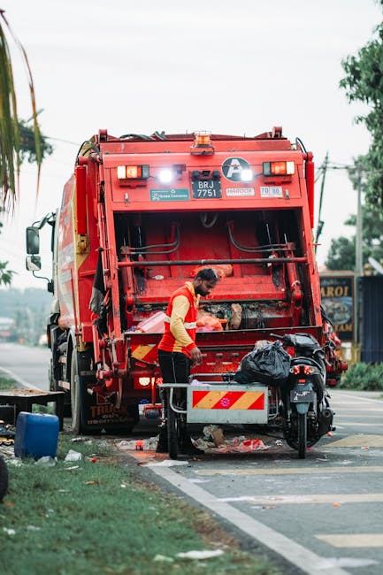 A waste collection worker in a yellow and red uniform is operating a red rubbish truck parked on the side of a urban street. The worker appears to be managing waste materials or debris, with the rear 