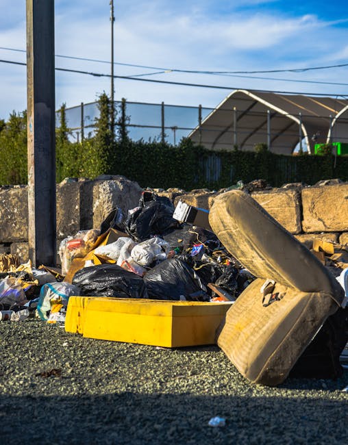 A waste collection worker in a yellow and red uniform is operating a red rubbish truck parked on the side of a urban street. The worker appears to be managing waste materials or debris, with the rear 